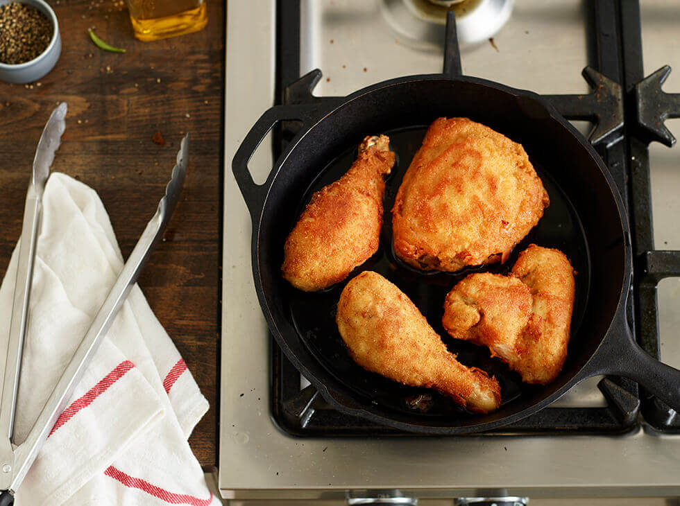 Frying chicken in a cast iron skillet on kitchen stove besides tongs and a dish towel